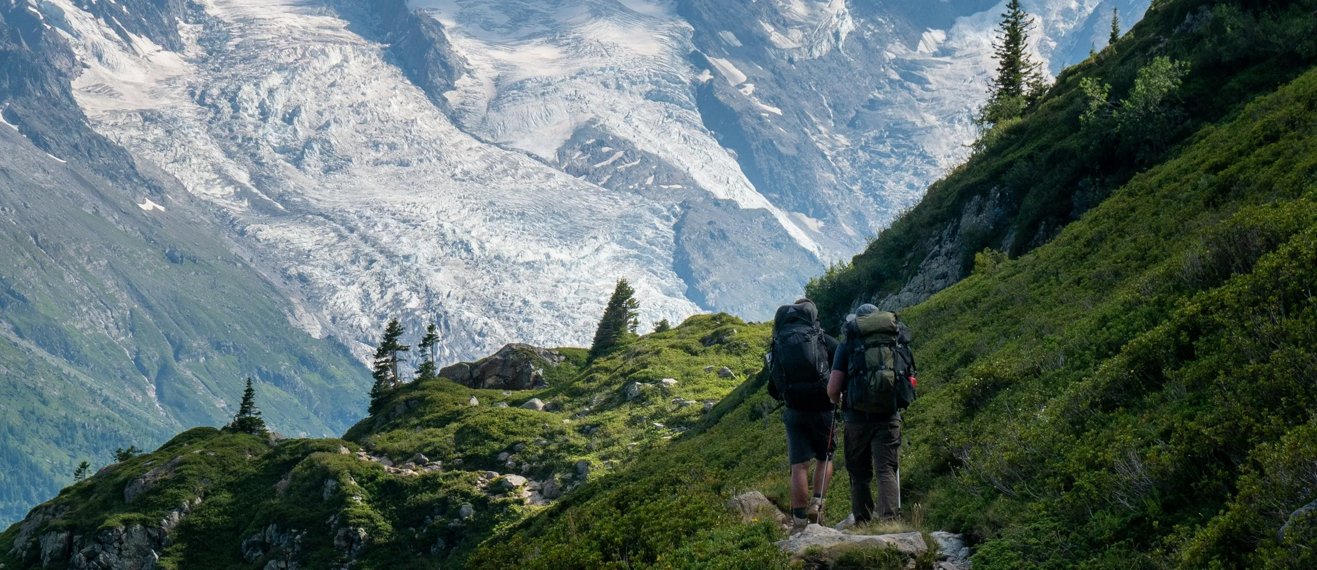 Two hikers on a trail above the Chamonix valley with the Bossons glacier behind
